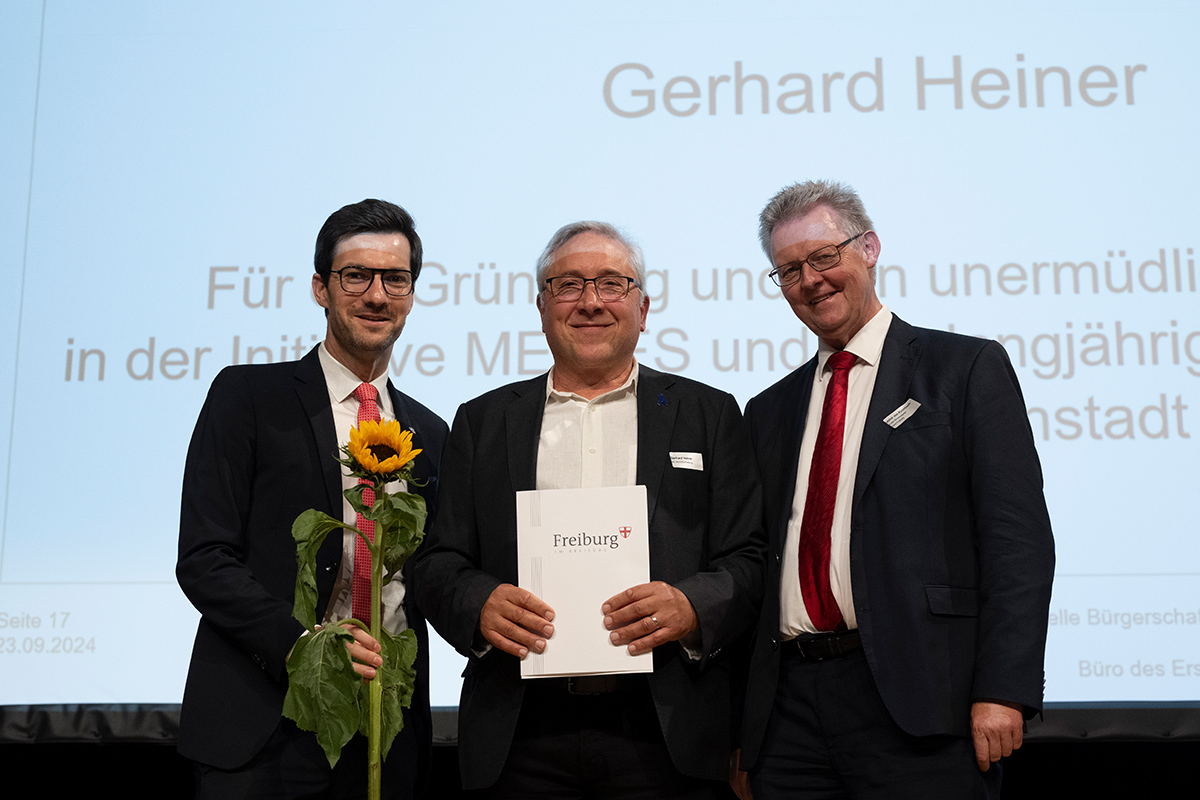 Bildbeschreibung: Foto von der Preisverleihung. Gerhard Heiner hält einen Umschlag mit der Aufschrift Freiburg. Neben ihm stehen der Oberbürgermeister Martin Horn mit einer Sonnenblume und der Erste Bürgermeister Ulrich von Kirchbach.