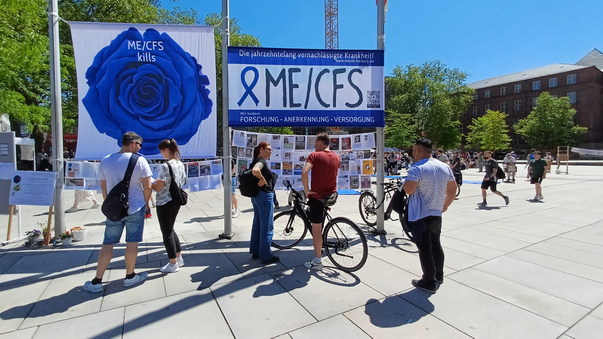 Foto: Passant*innen betrachten die Botschaften ME/CFS-Erkrankter. Über den Botschaften hängen zwei große Banner (links eine blaue Rose mit "ME/CFS kills", rechts "Die jahrzehntelang vernachlässigte Krankheit ME/CFS")